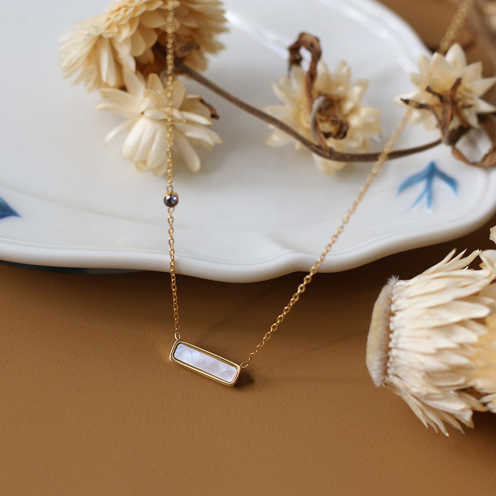 Gold necklace with a bar pendant on a brown surface with dried flowers.