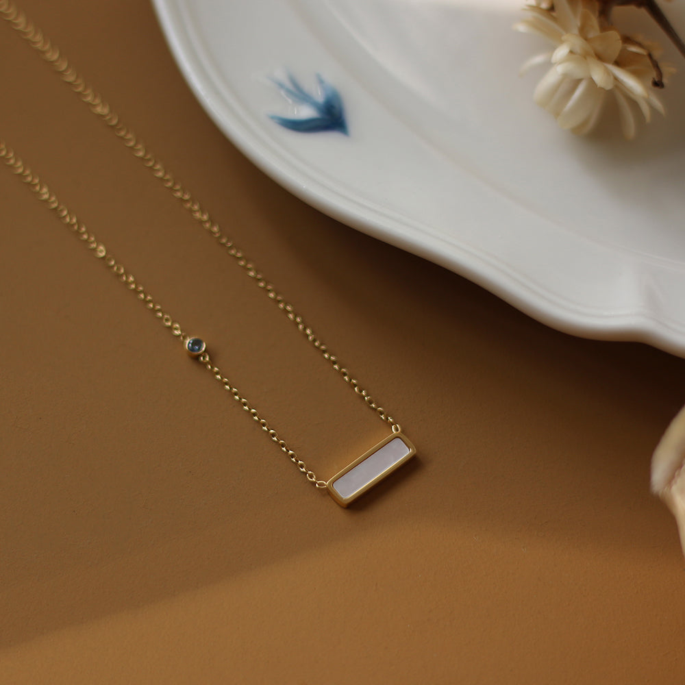 Gold necklace with a white pendant on a brown surface with a white plate and dried flowers in the background.