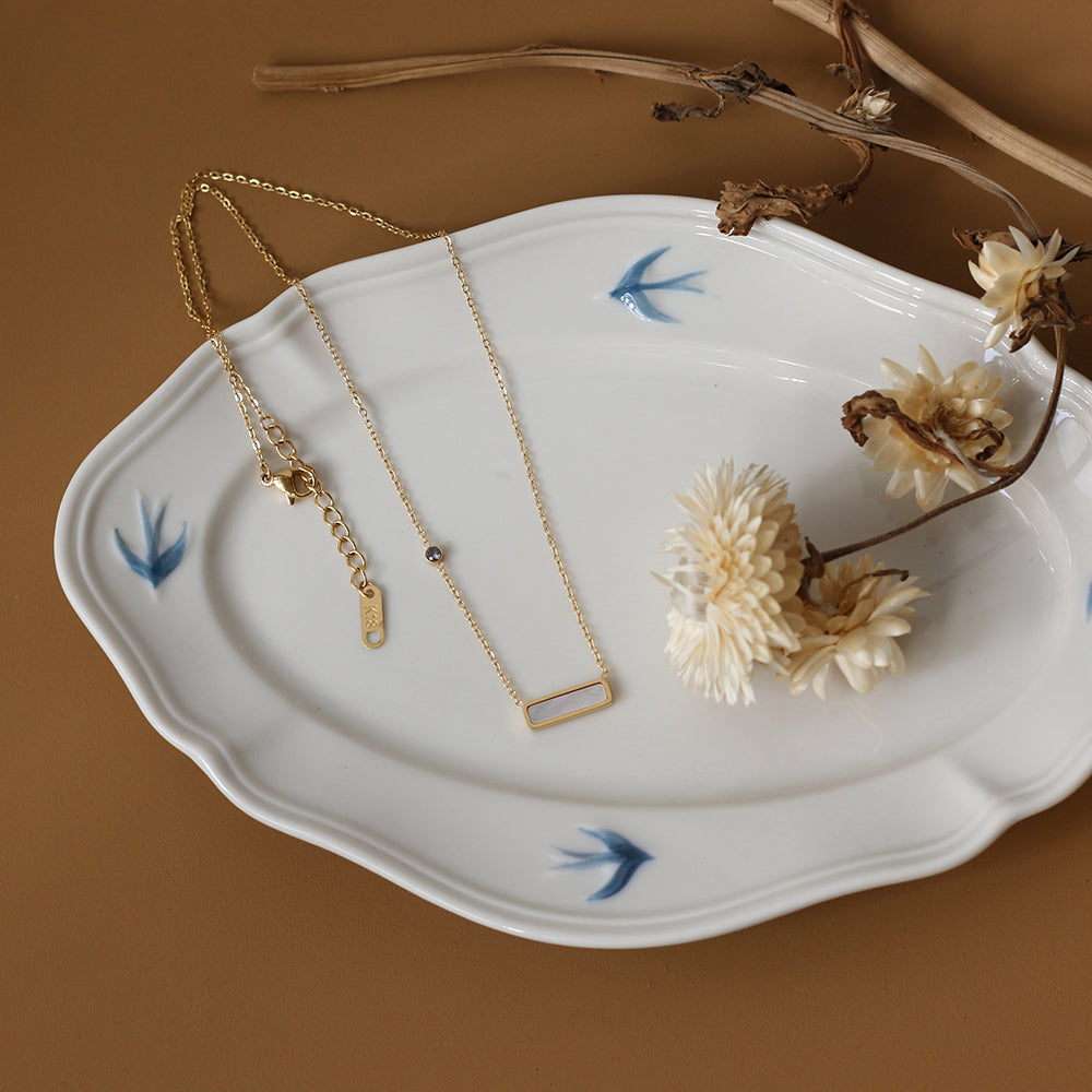 Gold necklace on a white dish with dried flowers and blue bird decorations against a brown background