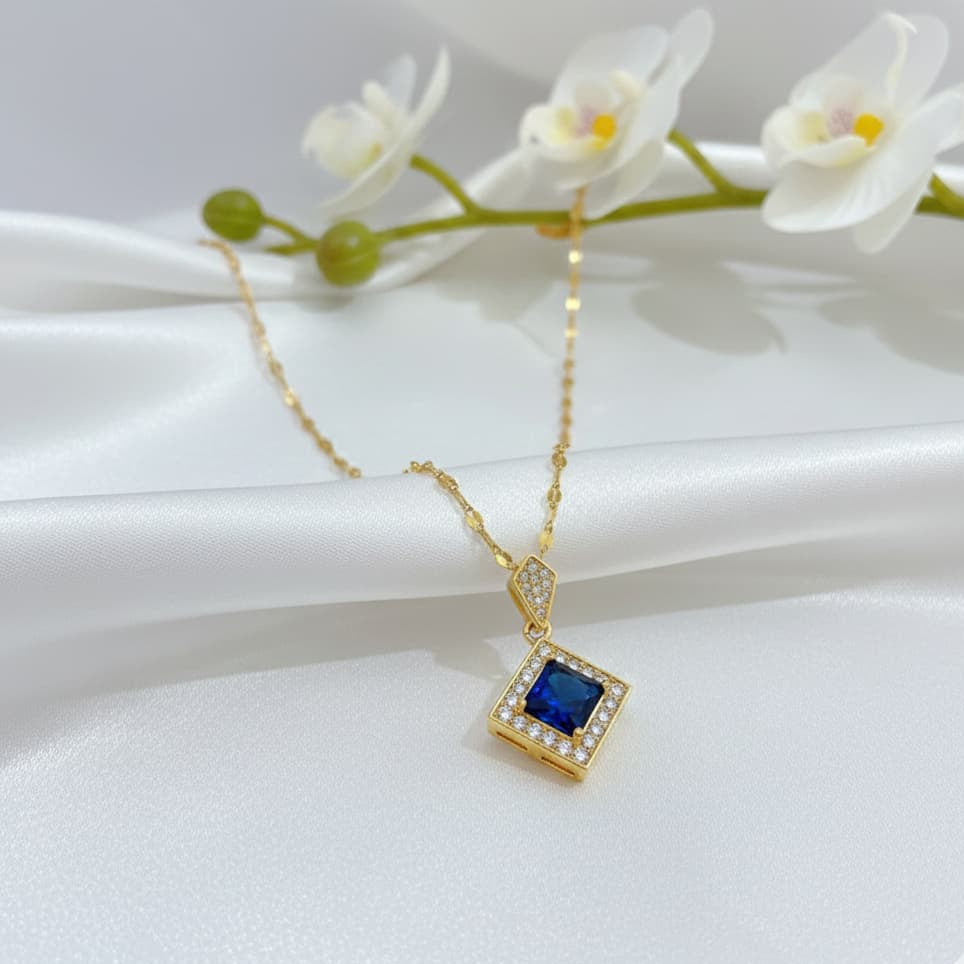 Gold necklace with a blue gemstone pendant on a white surface with flowers in the background