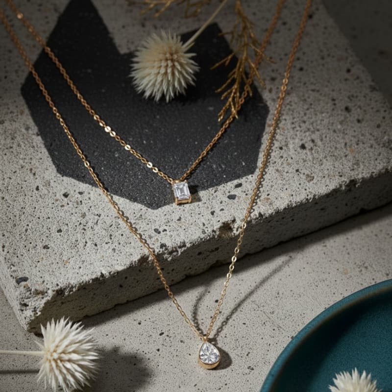 Two gold necklaces with diamond pendants on a textured surface with dried flowers.